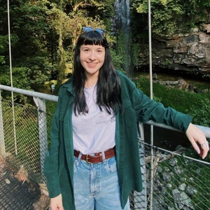 Photo of young woman in jeans, white t-shirt and green jacket smiling in front of waterfall. 