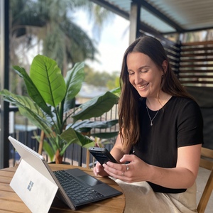 UNE graduate Becky Amon works on laptop and phone on verandah